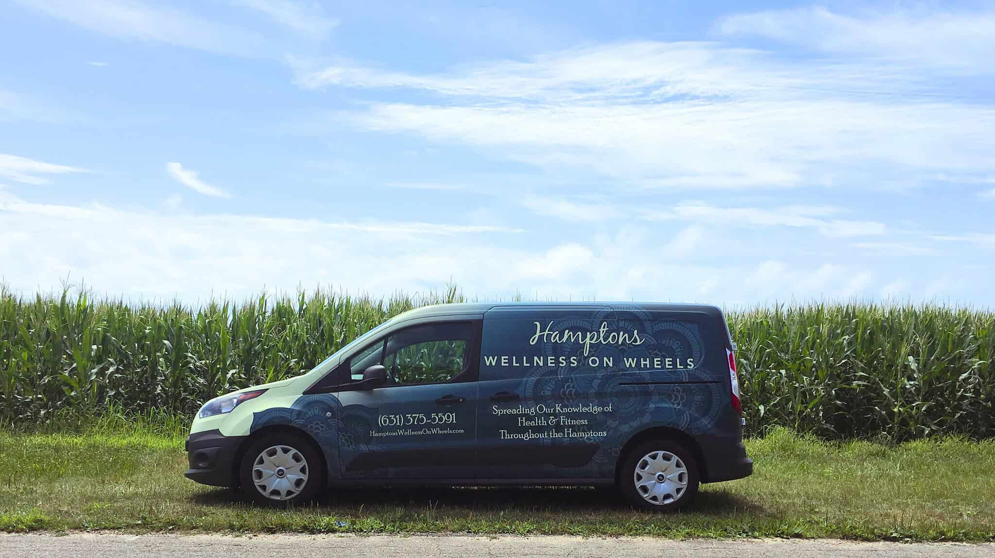 A green van with "Hamptons Wellness on Wheels" written on it is parked next to a field of tall corn. The van displays contact details and services offered.