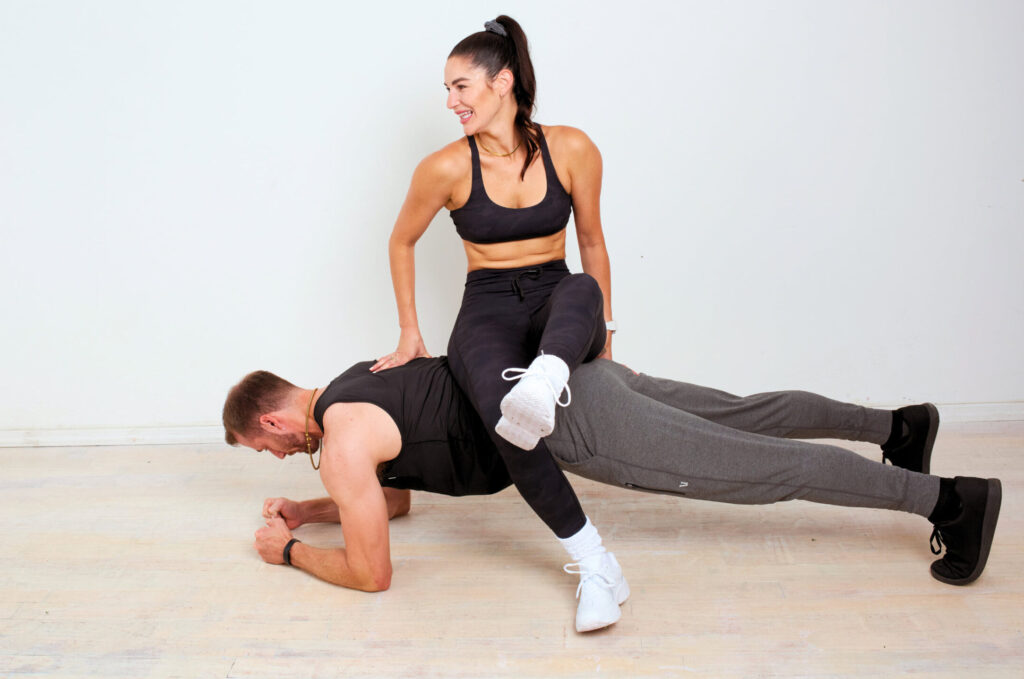A man performs a plank while a woman sits on his back, both in exercise attire, in a brightly lit room.