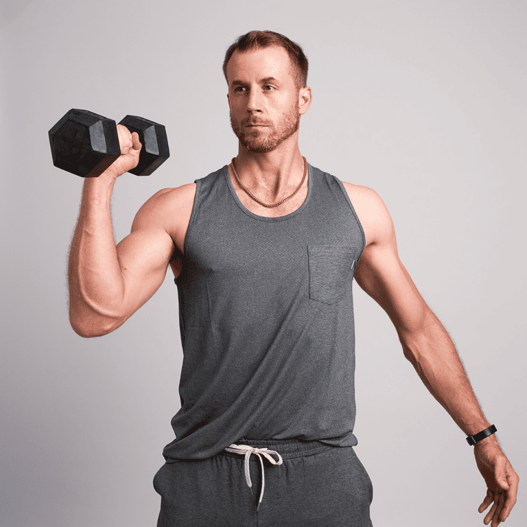 About a man in a gray tank top and sweatpants holding a dumbbell at shoulder height against a plain background.