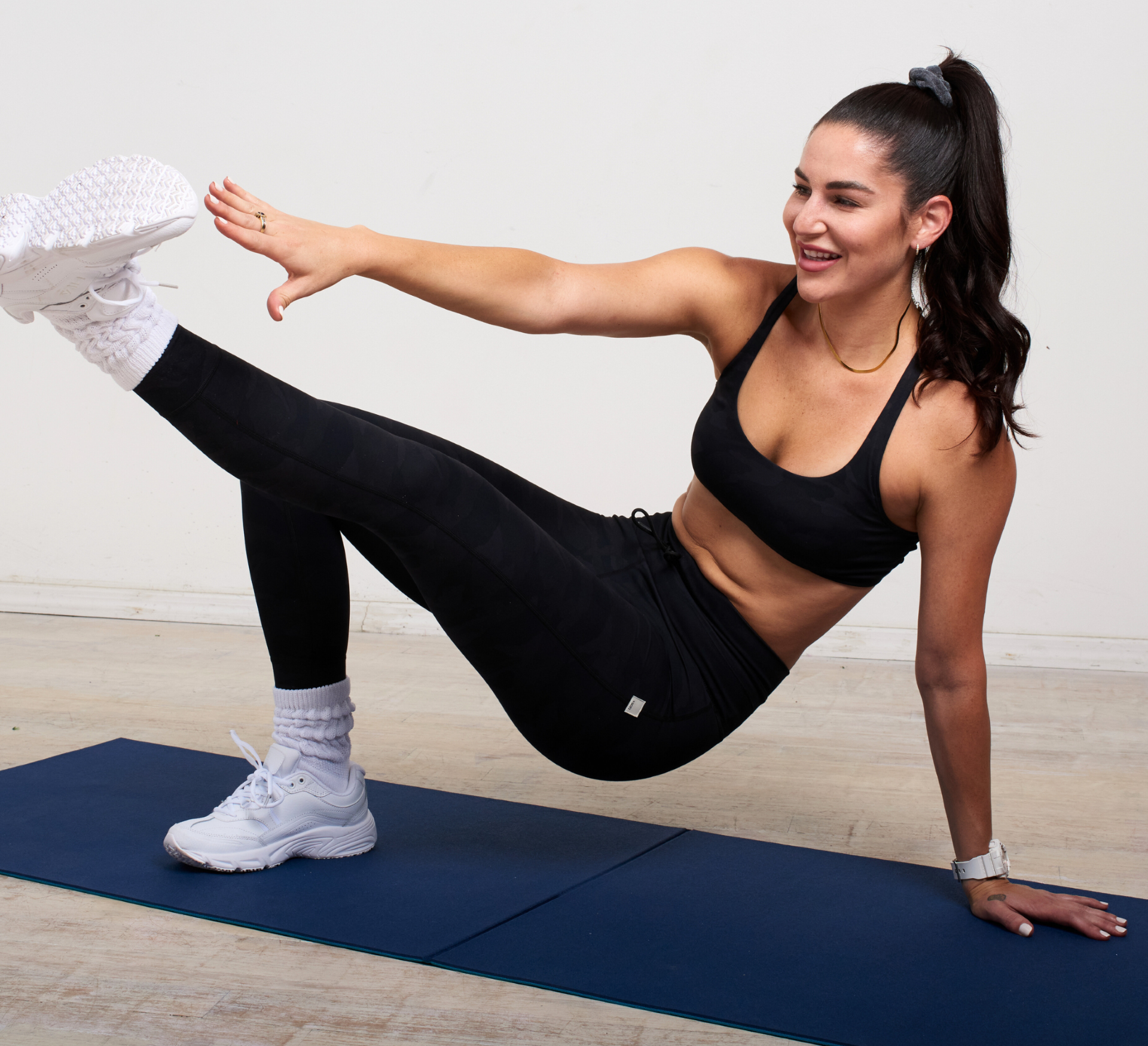 A woman in athletic attire performs a side plank with leg lift on a blue exercise mat. She extends her right arm and left leg, smiling as she balances on her right hand and left foot. This routine is all about mastering balance and strength together.