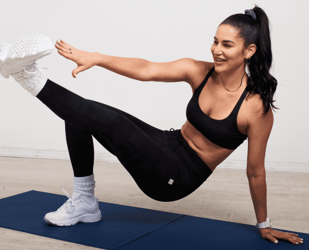 A woman in athletic attire performs a side plank with leg lift on a blue exercise mat. She extends her right arm and left leg, smiling as she balances on her right hand and left foot. This routine is all about mastering balance and strength together.