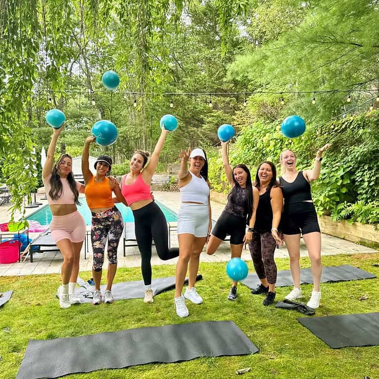 Seven women wearing sports attire stand on yoga mats outdoors, holding and tossing blue exercise balls under a leafy tree canopy. A pool is visible in the background.