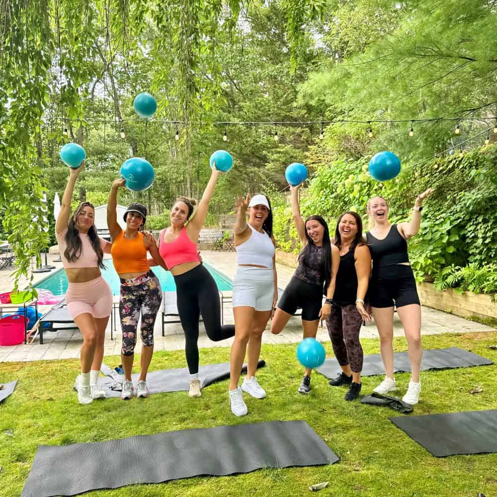 Seven women wearing sports attire stand on yoga mats outdoors, holding and tossing blue exercise balls under a leafy tree canopy. A pool is visible in the background.