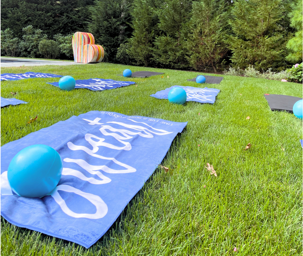 Colorful towels and blue exercise balls are arranged on a green lawn with trees and a multicolored chair in the background.
