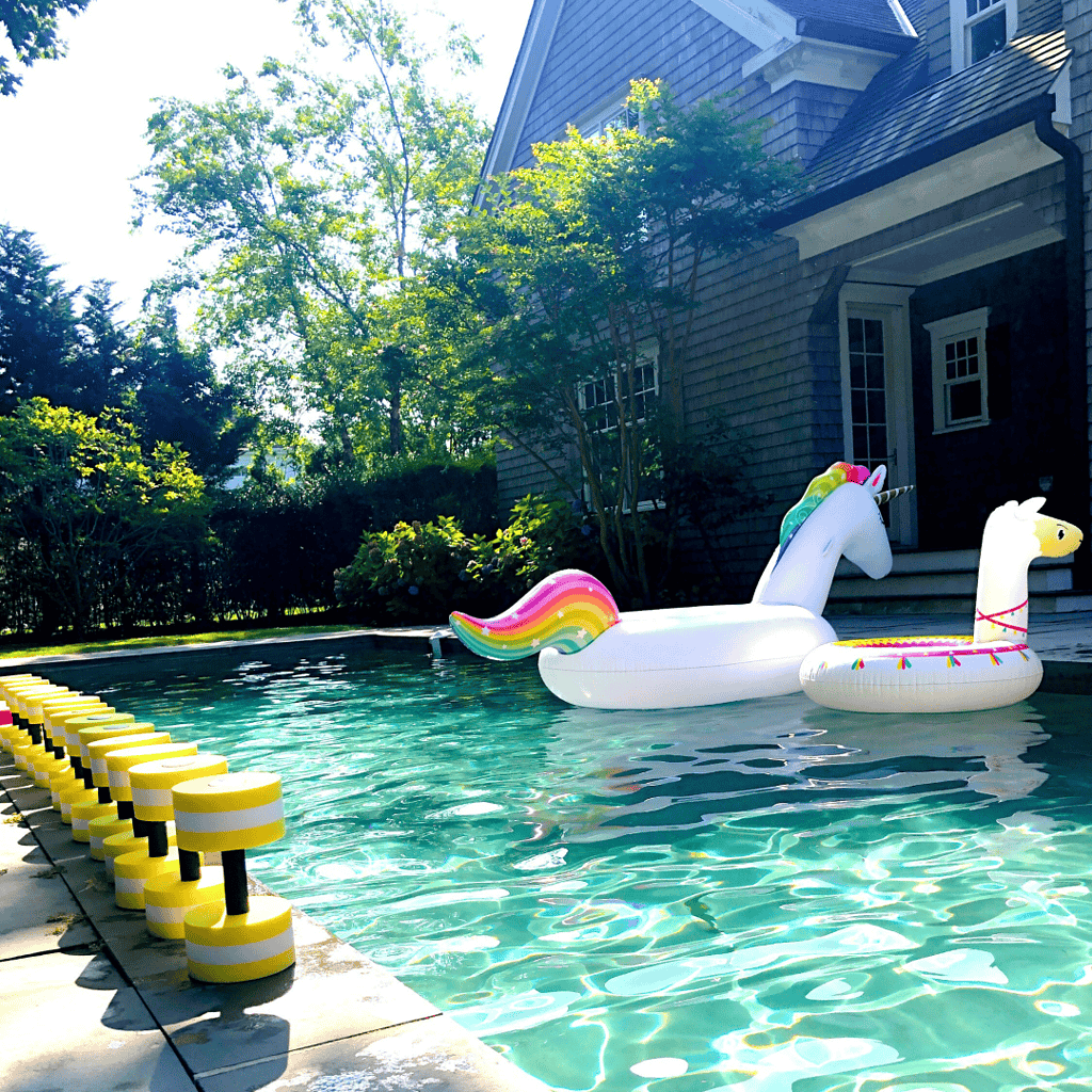 A swimming pool with floating inflatables shaped like a unicorn and a llama beside a house on a sunny day. Yellow and black segmented float markers line the pool's edge.