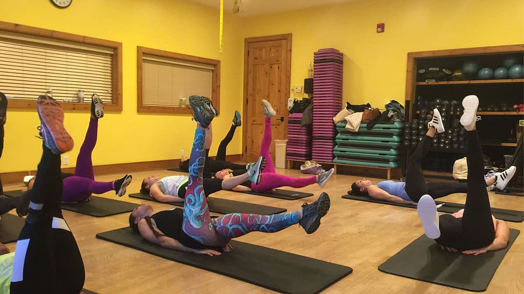 Six people on exercise mats in a room with yellow walls, performing leg lifts during a fitness class.