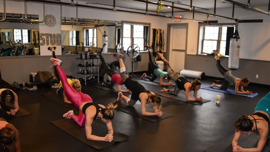 A group of people in a gym doing a fitness class, performing plank exercises with one leg lifted, while on yoga mats. Fitness equipment and mirrors are visible in the background.