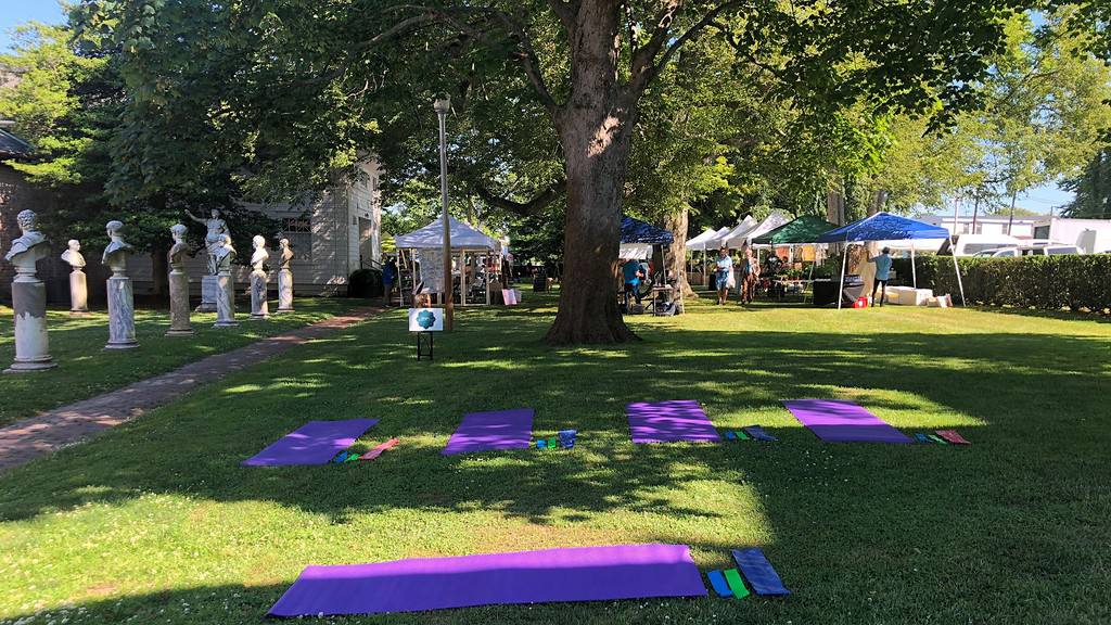 A grassy outdoor area with yoga mats laid out under a large tree. Nearby, there are white tents with people gathered, and a row of bust sculptures is visible on the left.