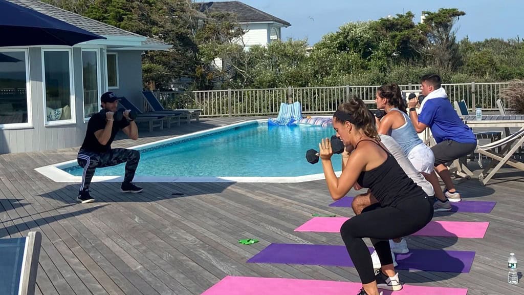 Four people doing a workout with dumbbells on yoga mats by an outdoor pool, led by an instructor.
