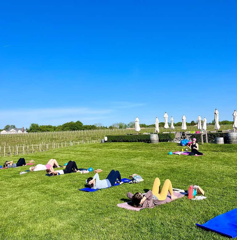 People are lying on yoga mats outdoors on a grassy field with a vineyard in the background under a clear blue sky.