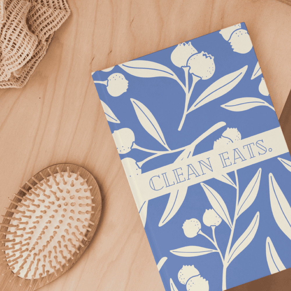 A book titled "Clean Eats" with a blue and white floral cover, available at our local bookstore, placed on a wooden surface next to a wooden hairbrush and a beige knitted cloth.