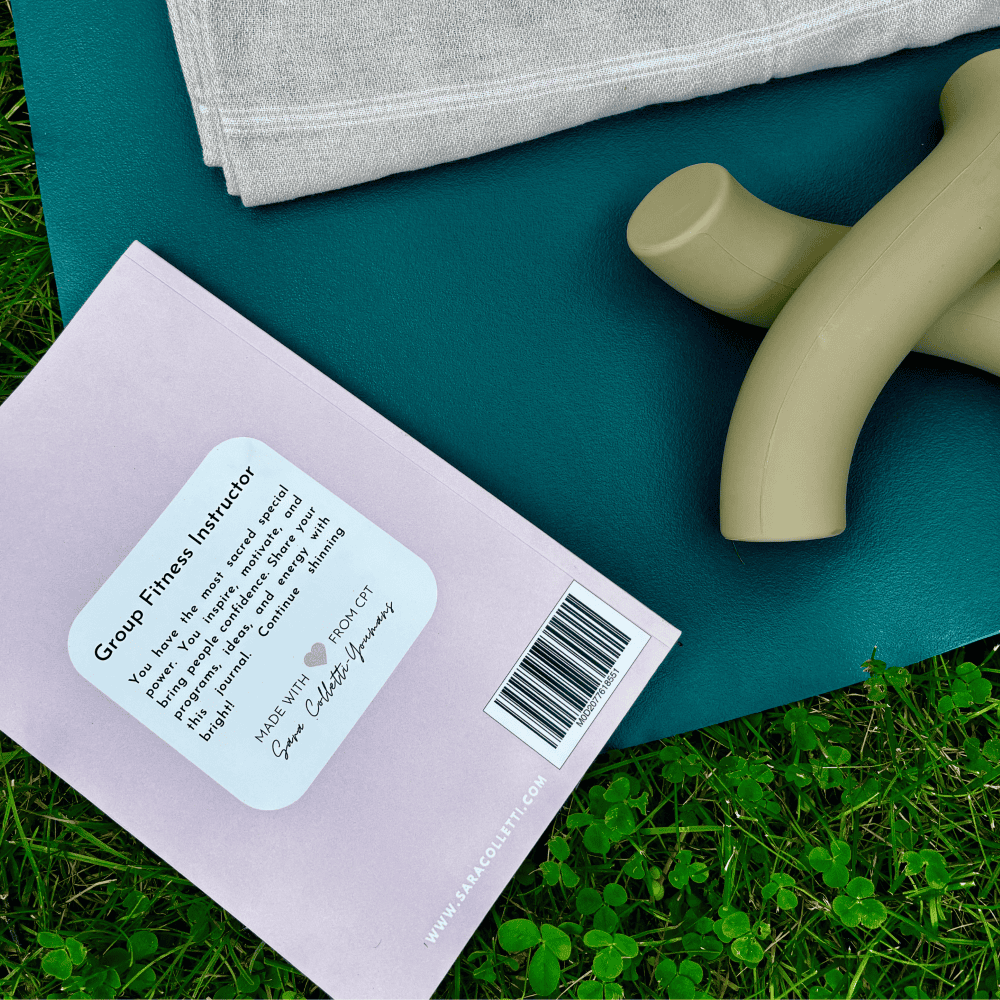 Close-up of a book titled "Group Fitness Instructor," available at Sara Colletti Bookstore, near a pair of beige hand weights and a folded towel on a blue yoga mat laid on green grass.