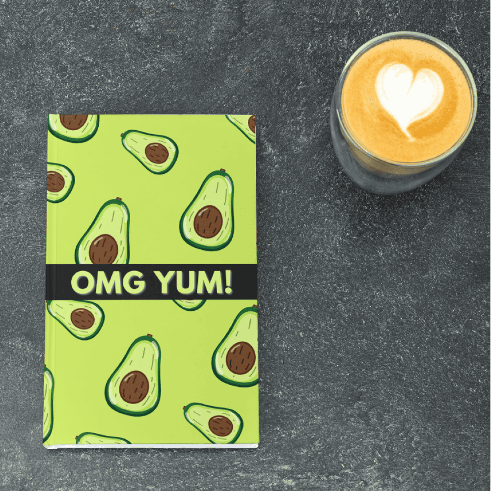 A green notebook with avocado illustrations and the text "OMG YUM!" lies next to a latte with a heart-shaped foam design on a dark textured surface, perfect for jotting down thoughts at your favorite bookstore.