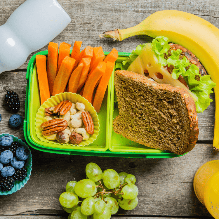 Lunchbox with a sandwich, carrot sticks, mixed nuts, and a banana on a wooden surface surrounded by grapes, blueberries, and blackberries. A white bottle and orange juice carton are also visible, providing a brain-boosting mix of nutrients.