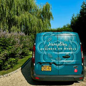How to start your own business. Rear view of a teal van with "Hamptons Wellness on Wheels" and a decorative pattern on the back, parked on a gravel driveway next to greenery and trees, offering mobile Pilates sessions.