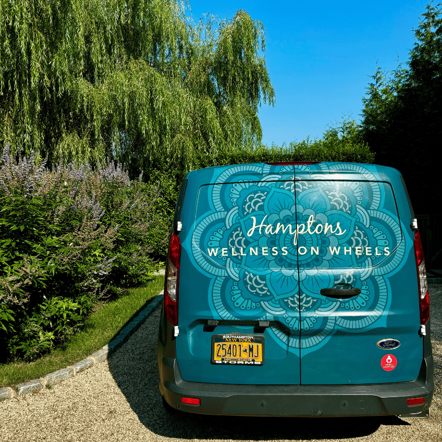 How to start your own business. Rear view of a teal van with "Hamptons Wellness on Wheels" and a decorative pattern on the back, parked on a gravel driveway next to greenery and trees, offering mobile Pilates sessions.