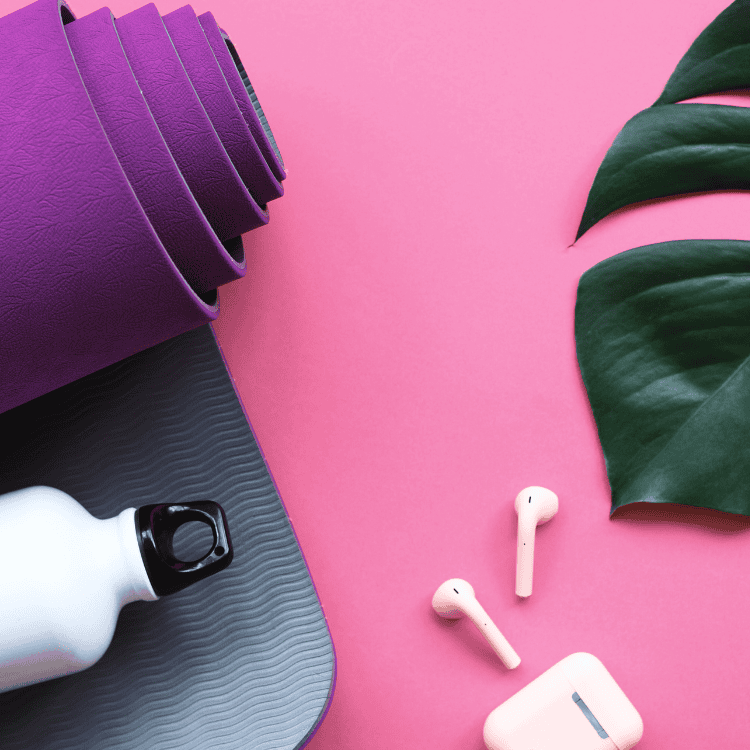 A photo of fitness trends that include rolled purple yoga mat, a white water bottle, wireless earbuds with a case, and green leaves on a pink background suggest tranquility before or after dinner.
