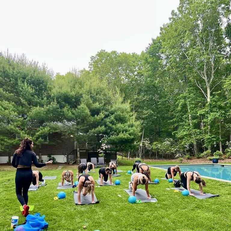 A group led by a Sara Colletti Group Fitness instructor exercises on yoga mats with small blue balls on a grassy lawn beside a swimming pool; trees and a house are in the background.