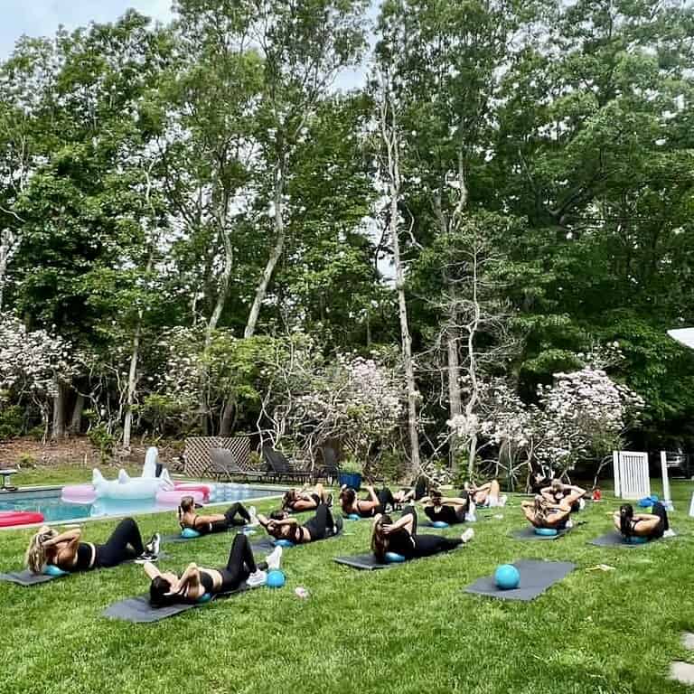A group of people in the Sara Colletti Group Fitness class exercise on mats with small balls on a grassy lawn, surrounded by trees and outdoor items.