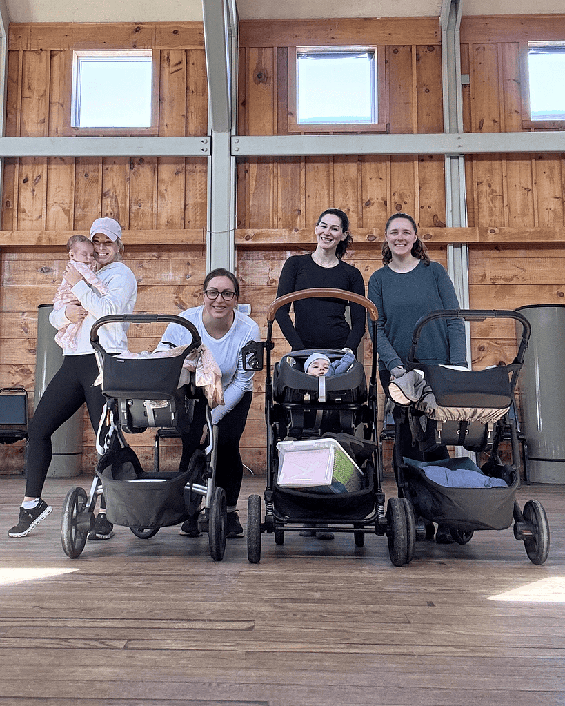 Four women stand indoors with three baby strollers; one woman is holding a baby. The background features wooden walls and natural light from upper windows.
