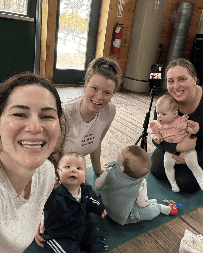 Three women and three babies are on a yoga mat indoors. One baby is sitting, two are crawling, and a phone on a tripod is set up nearby. There is a door and windows in the background.