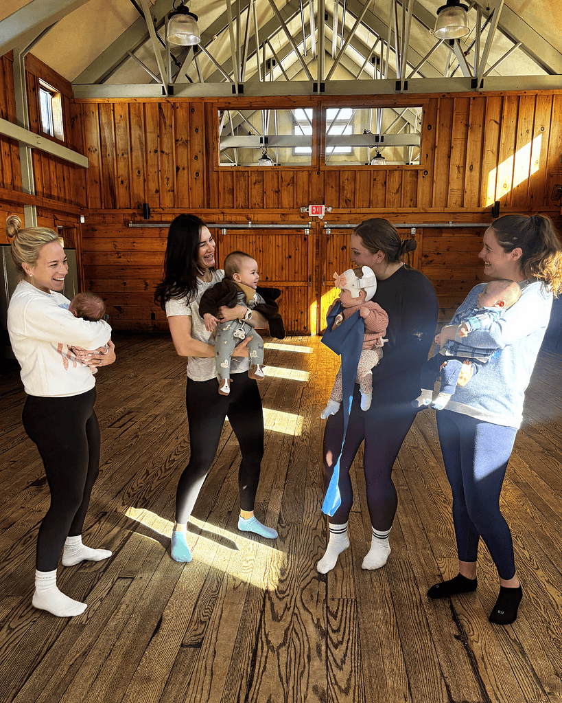 Five women stand in a sunlit wooden room, each holding a baby. They are smiling and appear to be having a friendly conversation.