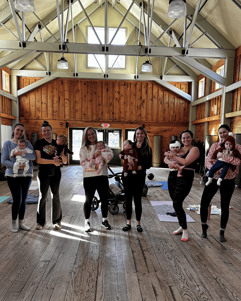 Seven women stand in a row inside a sunlit wooden hall, each holding a baby. Strollers and blankets are visible on the floor.