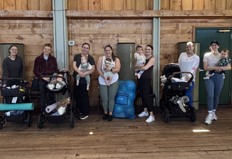 Seven women stand indoors, some holding babies and others with strollers, in front of a wooden wall scattered with bags and mats, participating in Sara Colletti Programs.