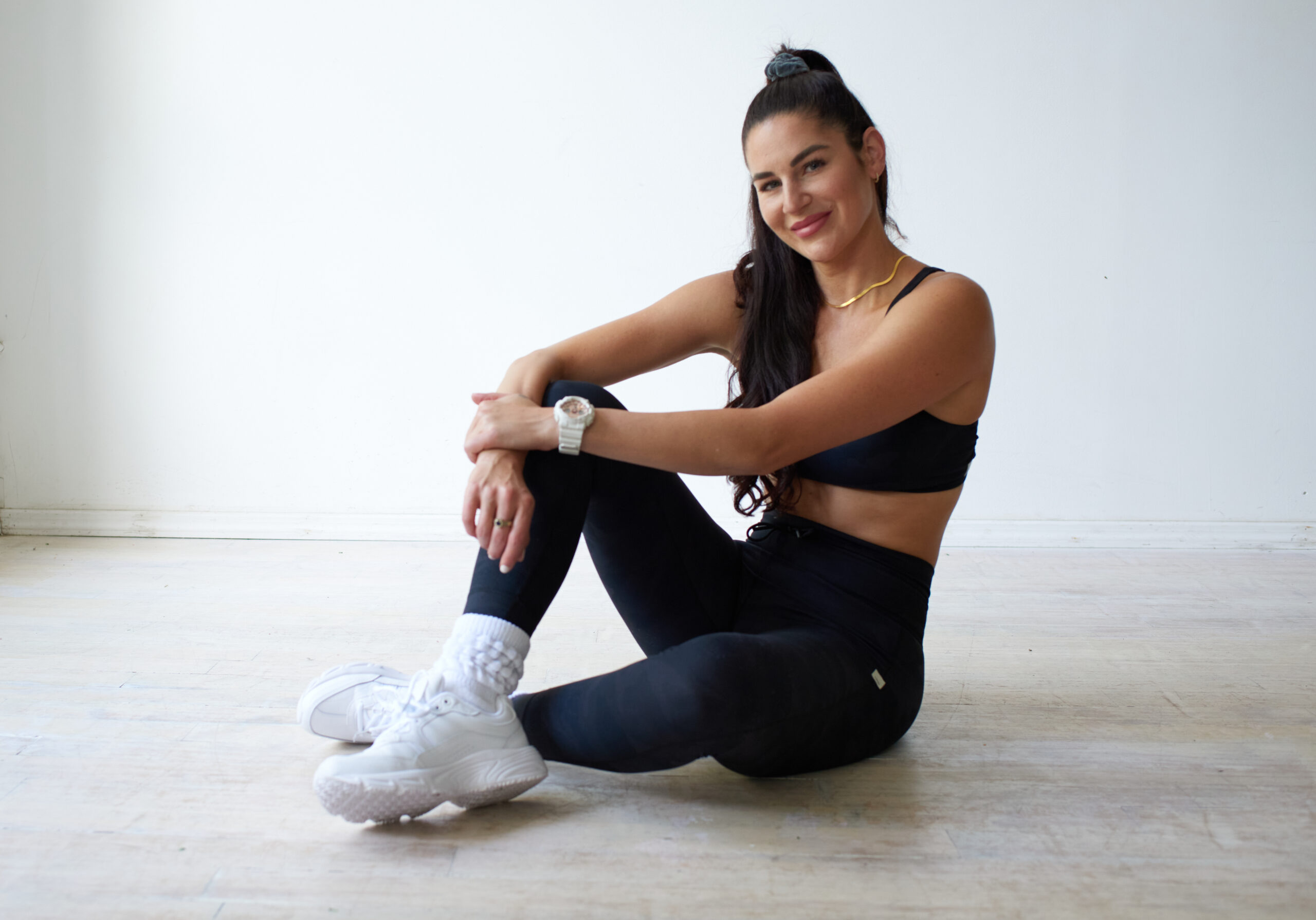 Sara in black workout clothes sits on the floor with legs crossed, smiling at the camera in a bright, minimal room—capturing the welcoming spirit of Sara Colletti Personal Fitness.