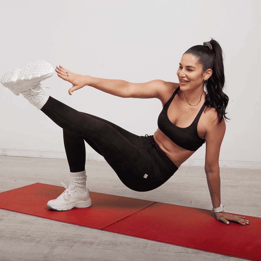 Woman in black workout clothes balances on one hand and foot on a red exercise mat, raising her opposite arm and leg, in a fitness studio with a white wall and light wood floor—a scene that perfectly reflects the mindful movement approach of Sara Colletti. About dedication and strength, this pose exemplifies both balance and grace.