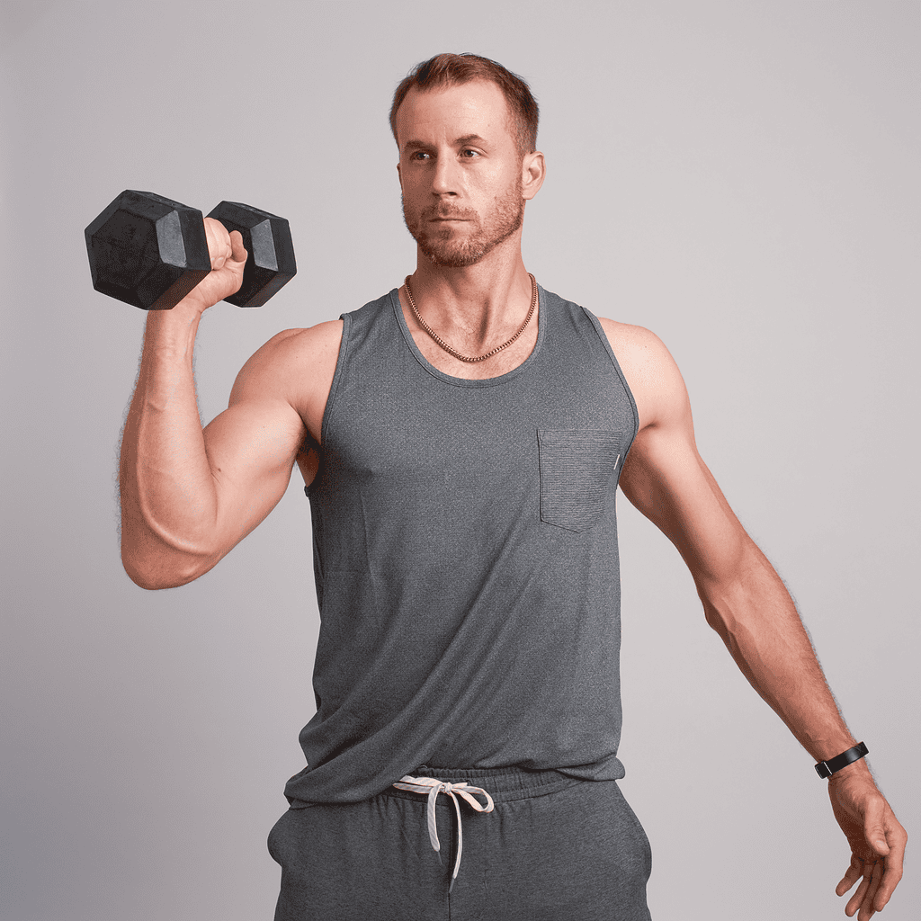 A man wearing a gray tank top and sweatpants holds a dumbbell at shoulder height with his right hand against a plain background, embodying the active lifestyle celebrated in Sara Colletti About.